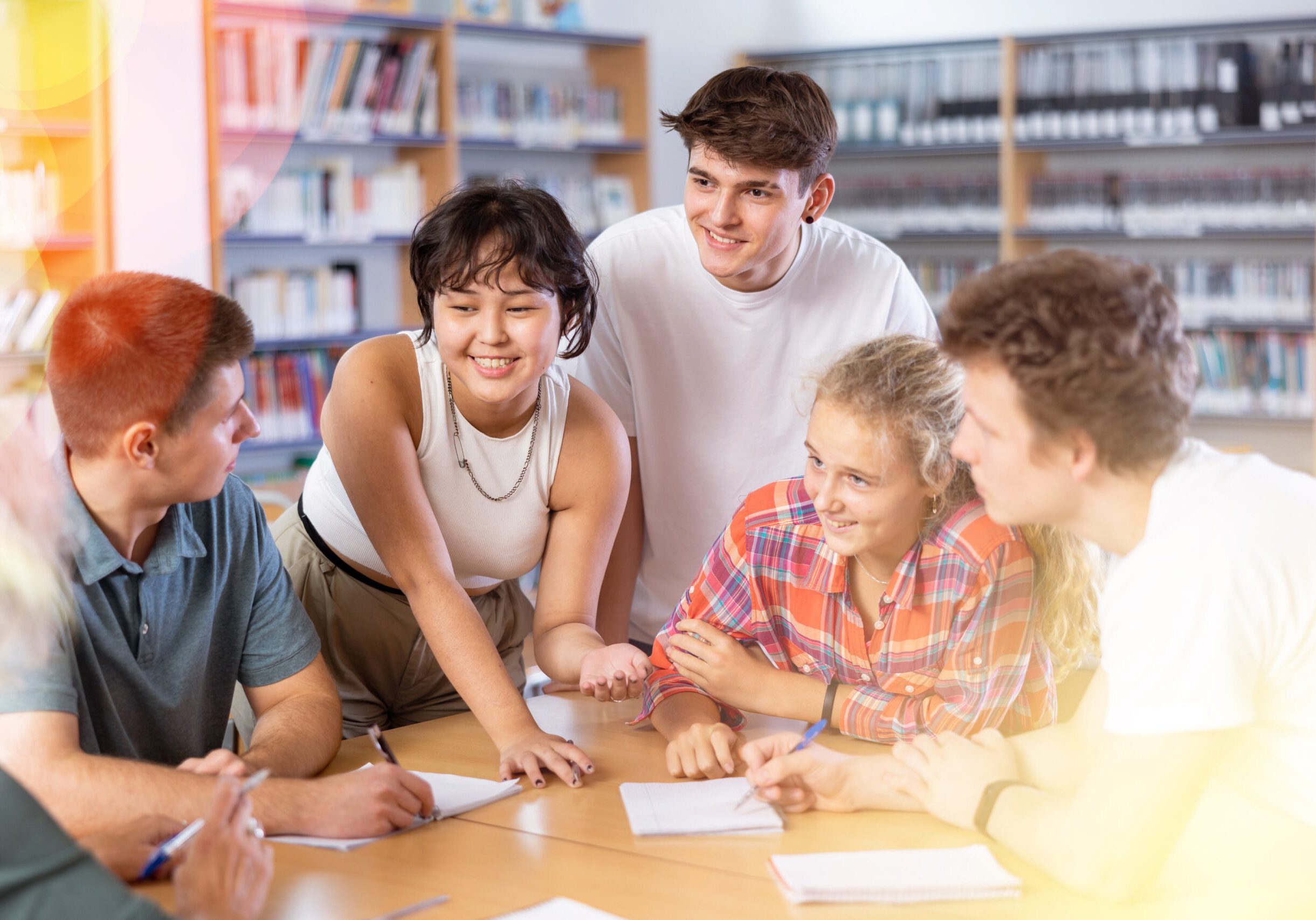 Group of fifteen-year-old schoolchildren are discussing something and making notes in copybooks, preparing for classes in the school library